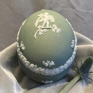 May include: A pale green egg-shaped decorative object with white relief detailing. The design includes a cherub figure and floral garlands. The object is resting on a soft, gray surface, with a thistle flower in the foreground.