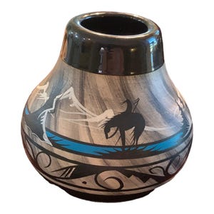 May include: A decorative ceramic vase with a black rim and a gray and white body. The vase features a Native American design with a black silhouette of a person on a horse, mountains, and a blue river.