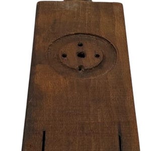 May include: A vintage wooden butter mold with a handle. The rectangular mold has a circular design on the top and two vertical slots at the bottom. The wood has a dark brown color and shows signs of wear.