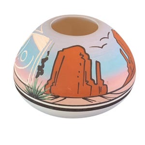 May include: A small, round, ceramic vase with a wide opening. The vase features a landscape design with a large orange rock formation, blue sky, and desert plants. The base is white with black and pink stripes.