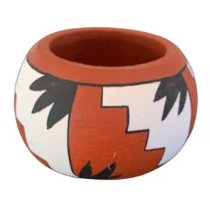 May include: A small, round, hand-painted ceramic pot with a wide opening. The pot features a design of alternating white, black, and orange geometric shapes. The rim and interior are orange. This decorative piece could be used as a small planter or a decorative bowl.