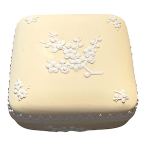May include: A square, cream-colored trinket box with a white floral design on the lid. The box has a decorative border and a raised floral branch in the center. The box is likely made of ceramic or porcelain.