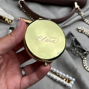May include: A gold-colored, round compact mirror with the name "Clara" in cursive script. The mirror is held in a hand, with a purse, hair clips, and a chain in the background. The compact is a beauty accessory.