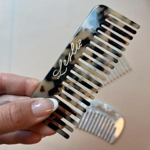 May include: A tortoiseshell-patterned comb with the name "Leslie" in gold script. The comb has evenly spaced teeth and is held in a hand. Another comb is visible in the background.