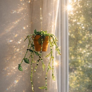 Op de afbeelding: Een hangende terracotta pot met een kralen plantontwerp. De plant heeft groene en olijfkleurige kralen, met bladvormige accenten. De pot hangt aan een delicate ketting met kristalachtige kralen, bij een raam met zonlicht.