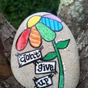 May include: A painted stone with a colorful flower design and the words "don't give up." The flower has red, orange, yellow, green, and blue petals. The stem and leaves are green, and the background is a natural stone color.