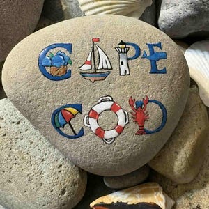 Peut inclure: Une pierre peinte avec les mots "CAPE COD" en lettres bleues. Les lettres sont décorées d'images côtières : un voilier, un phare, un parapluie, une bouée de sauvetage et un homard. La pierre est entourée d'autres pierres et coquillages.