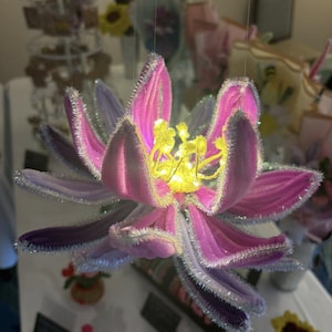 Könnte beinhalten: Eine große, dekorative Blume mit rosa und lila Blütenblättern, akzentuiert mit silbernem Glitzer und einem gelben Zentrum. Die Blume hängt an einem Faden und zeigt ihr kompliziertes Design und ihre leuchtenden Farben. Die Blütenblätter haben eine weiche, samtige Textur.
