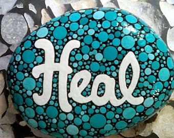 May include: A painted stone with the word "Heal" in white script. The stone is covered in teal and turquoise polka dots of varying sizes against a black background. The stone is oval-shaped and sits on a bed of other stones.