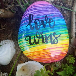 Puede incluir: Una piedra pintada con un diseño de rayas de arcoíris y las palabras "love wins" en escritura negra. La piedra está rodeada de arena, conchas y vegetación.
