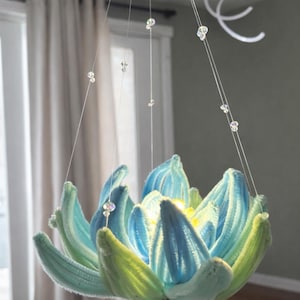 Op de afbeelding: Een handgemaakte kroonluchter in de vorm van een lotusbloem. De bloemblaadjes zijn een mix van blauw en groen, met een zachte, getextureerde afwerking. Heldere kristallen hangen aan de armatuur en voegen glans toe. Een decoratieve verlichtingsarmatuur.