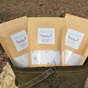 May include: Three brown paper bags filled with white bath salts. Each bag has a label with the text "Compass Ranch Apothecary" and a floral design. The bags are sitting in a green metal bucket.