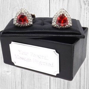 May include: A pair of silver cufflinks with a red gemstone in the centre, surrounded by smaller clear gemstones. The cufflinks are in a black box with a silver plaque that says "Your Personal Message Engraved".