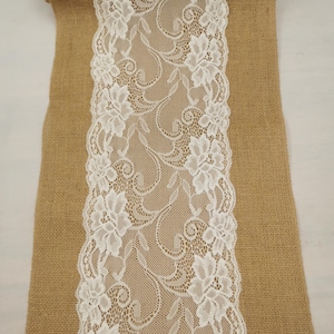 Op de afbeelding: Een tafelloper van beige jute met een brede, centrale strook witte bloemenkant. Het kant heeft een gedetailleerd, ingewikkeld patroon van bloemen en krullende ranken dat verticaal door het midden van het bruine, getextureerde materiaal loopt.