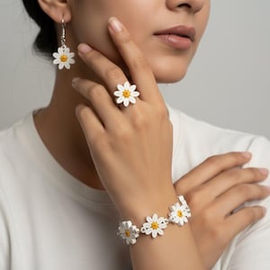May include: White daisy flower jewelry set including earrings, a ring, and a bracelet. The daisy petals are white with yellow centers. The jewelry is worn on a person's hand and wrist.