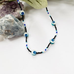 May include: A beaded necklace featuring small black, white, and blue seed beads interspersed with five round, light blue clay beads. Each clay bead displays a dark blue graphic of a creature with a small white star above its head.