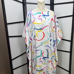 May include: A white, flowing caftan dress with a colorful abstract design. The dress features a pattern of curved lines in red, yellow, blue, green, and purple. The dress is displayed on a mannequin against a black and white folding screen.