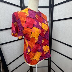 May include: A short-sleeved blouse with a vibrant geometric pattern in shades of orange, red, yellow, and purple. The blouse is displayed on a mannequin, showcasing its design and fit. The fabric appears to be lightweight.