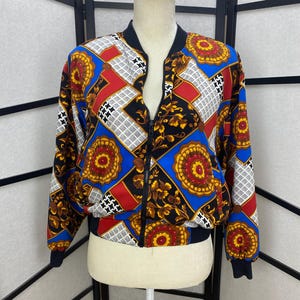 May include: A black bomber jacket with a colorful geometric pattern. The pattern features red, blue, gold, and white colors. The jacket has a zipper closure and a ribbed collar, cuffs, and hem.
