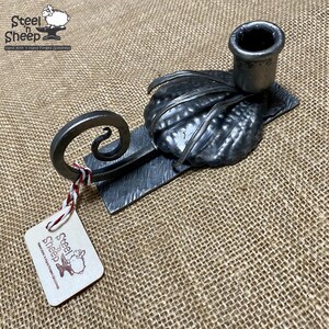 May include: A hand-forged metal candle holder with a leaf design and a scroll-shaped hook. The holder is made by Steel 'n Sheep, a company that specializes in hand-crafted goods.