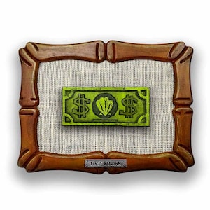May include: A framed artwork featuring a green dollar bill with dollar signs and a central design. The artwork is set against a textured, off-white background within a brown, ornate frame. A small metal plate reads "FIRST DOLLAR".