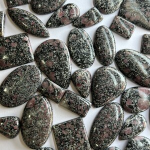 Op de afbeelding: Een verzameling van gepolijste, zwart en roze gevlekte edelsteen cabochons in verschillende maten en vormen. De cabochons zijn gerangschikt op een wit oppervlak.