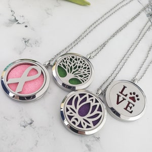 Peut inclure: Quatre colliers d'aromathérapie argentés avec des motifs différents. Les motifs comprennent un ruban rose, un arbre vert, une fleur de lotus violette et une empreinte de patte argentée avec le mot "LOVE".