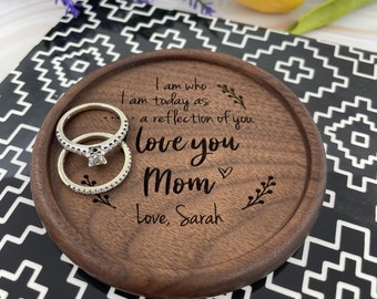 May include: A wooden ring dish with the inscription "I am who I am today as a reflection of you love you Mom Love, Sarah" and two rings on top.
