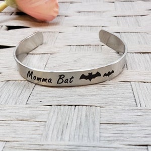 May include: A silver bangle bracelet with the words "Momma Bat" engraved on it. Two bat silhouettes are also engraved on the bracelet.