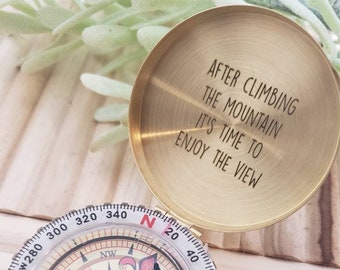 May include: A brass compass with a white face and black needle. The compass has a lid with an engraved message: "After climbing the mountain it's time to enjoy the view."