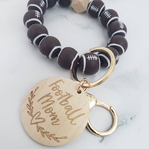 Op de afbeelding: Een bruine en witte voetbal-sleutelhanger met een houten hanger waarop "Football Mom" staat met een hartje en bladeren. De sleutelhanger is gemaakt van bruine siliconen kralen met witte voetbalontwerpen.