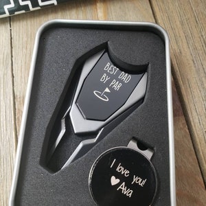 May include: A silver golf ball marker with the text "Best Dad By Par" and a golf flag icon. A black golf ball marker with the text "I love you! Ava" and a heart icon is also included. The markers are in a silver tin.