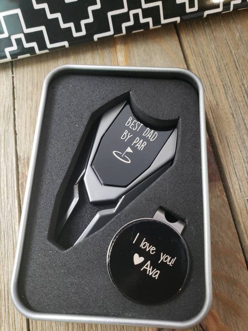 May include: A silver golf ball marker with the text "Best Dad By Par" and a golf flag icon. A black golf ball marker with the text "I love you! Ava" and a heart icon is also included. The markers are in a silver tin.