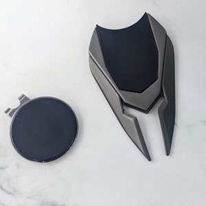 May include: A black and silver golf ball marker with a geometric design. The marker has a clip on the back to attach to a golf bag.