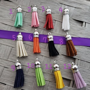 May include: Twelve different colored tassels with silver tops. The tassels are made of suede and are hanging from a woven surface.