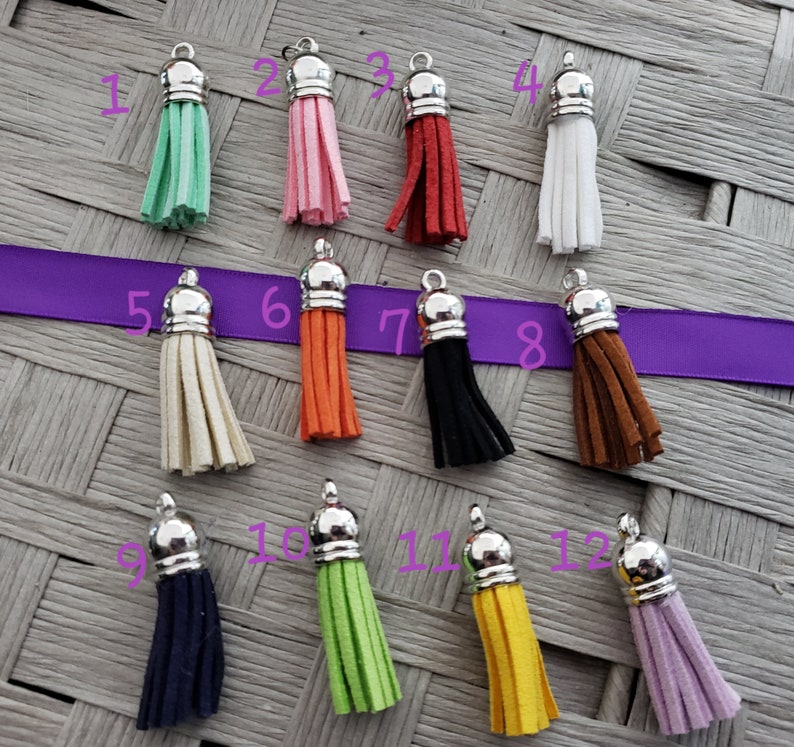 May include: Twelve different colored tassels with silver tops. The tassels are made of suede and are hanging from a woven surface.