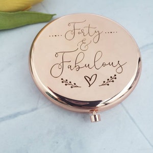 May include: A rose gold compact mirror with the words "...Forty & Fabulous..." engraved on the front. The mirror has a small heart and floral designs around the text.