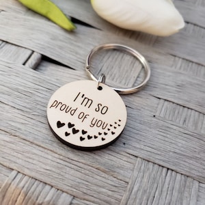 May include: Wooden keychain with a round tag that says "I'm so proud of you" with small hearts around the edge.