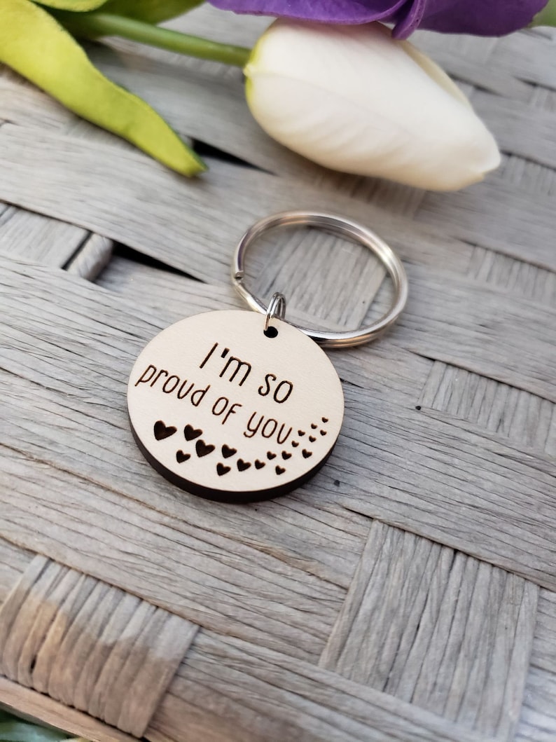 May include: Wooden keychain with a round tag that says "I'm so proud of you" with small hearts around the edge.