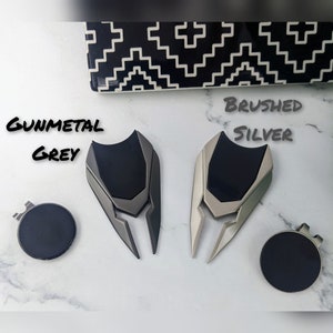 May include: Two golf ball divot tools, one in gunmetal grey and one in brushed silver. Both tools have a black rubber grip and a sharp, pointed end for fixing divots on the golf course.