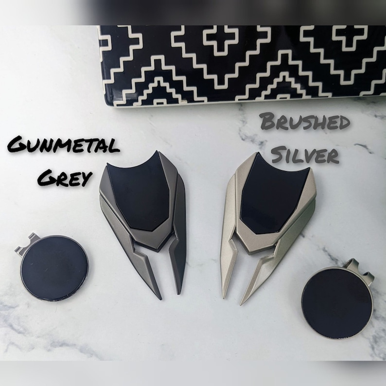May include: Two golf ball divot tools, one in gunmetal grey and one in brushed silver. Both tools have a black rubber grip and a sharp, pointed end for fixing divots on the golf course.