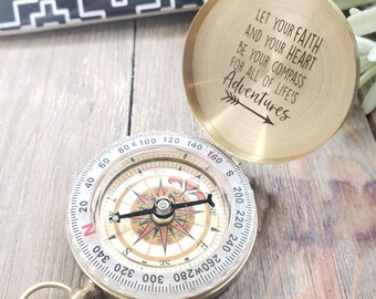 May include: A brass compass with a quote engraved on the top: "Let your faith and your heart be your compass for all of life's adventures". The compass has a white face with black markings and a red needle.