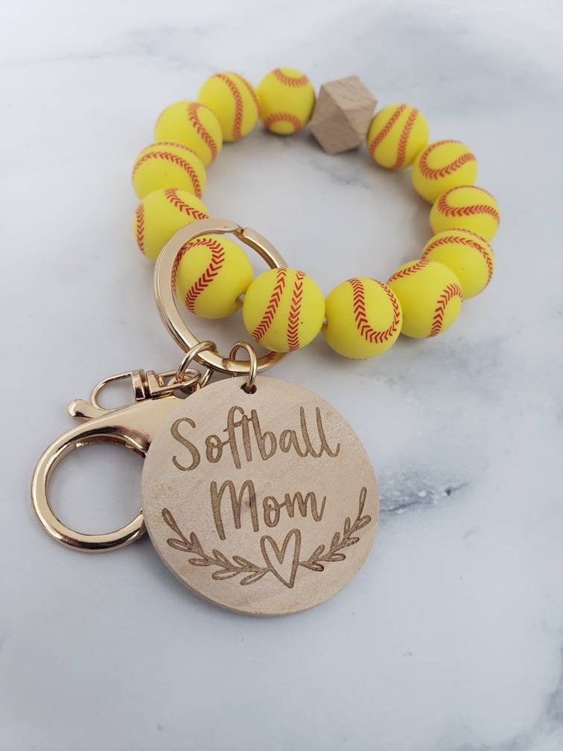 Op de afbeelding: Een gele en rode softball sleutelhanger met een houten hanger waarop "Softball Mom" staat met een hartje en bladeren.
