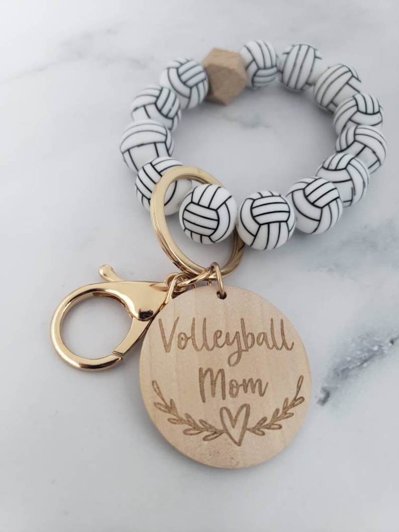 Op de afbeelding: Een wit-zwarte volleybal-sleutelhanger met een houten hanger waarop "Volleyball Mom" met een hart staat. De sleutelhanger is gemaakt van een kralenarmband met een gouden sluiting.