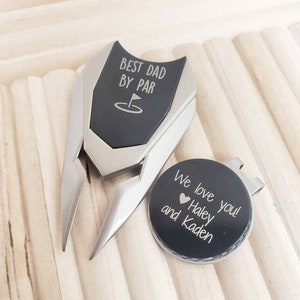 May include: A black golf ball marker with the text "We love you! Haley and Kaden" and a small heart. The marker is attached to a silver golf ball retriever with a black handle that says "Best Dad By Par" with a golf hole icon.
