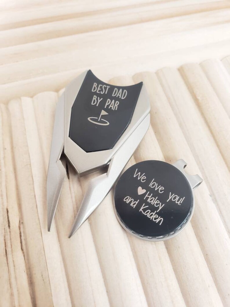 May include: A black golf ball marker with the text "We love you! Haley and Kaden" and a small heart. The marker is attached to a silver golf ball retriever with a black handle that says "Best Dad By Par" with a golf hole icon.