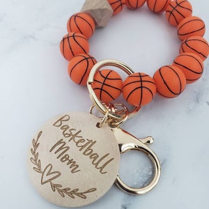Op de afbeelding: Een houten sleutelhanger met een basketbalthema. De sleutelhanger heeft een ronde houten hanger met de tekst "Basketball Mom" en een hartje. De hanger is bevestigd aan een gouden metalen sluiting en een armband gemaakt van oranje basketbalvormige kralen.
