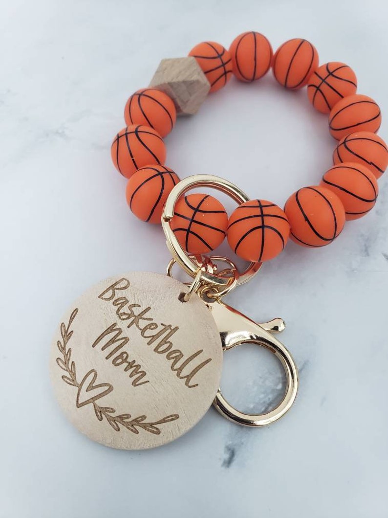 Op de afbeelding: Een houten sleutelhanger met een basketbalthema. De sleutelhanger heeft een ronde houten hanger met de tekst "Basketball Mom" en een hartje. De hanger is bevestigd aan een gouden metalen sluiting en een armband gemaakt van oranje basketbalvormige kralen.