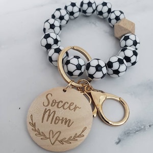 Op de afbeelding: Een sleutelhanger met een zwart-witte voetbalbedel en een houten tag met de tekst "Soccer Mom" met een hartje en bladeren.