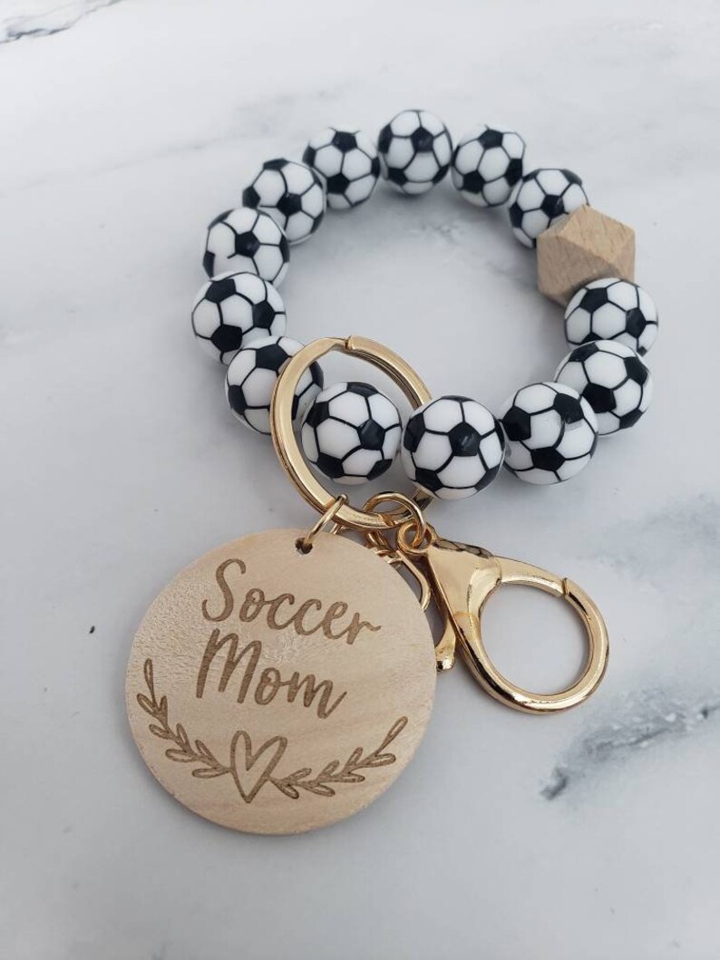 Op de afbeelding: Een sleutelhanger met een zwart-witte voetbalbedel en een houten tag met de tekst "Soccer Mom" met een hartje en bladeren.